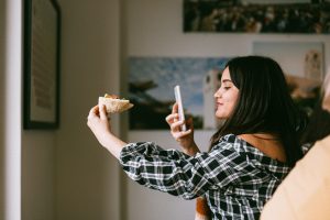 woman poses with food in her hand, taking a photo with a smartphone